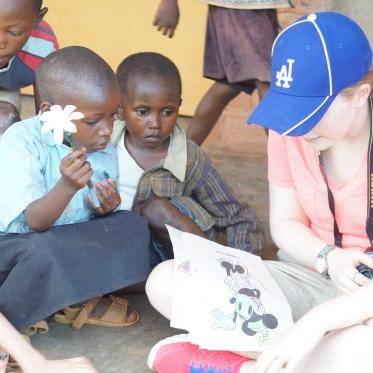 一位戴着蓝色棒球帽的妇女给坐在她周围感兴趣的孩子们读书. One holds a flower.