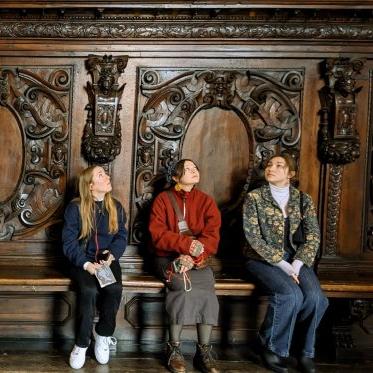 3名学生坐在长凳上，仰望着一堵雕刻精美的木墙.